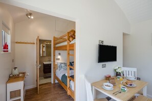 A bedroom with a bunk bed, a bedside table with a lamp, a TV mounted on the wall, and a breakfast table with food and drinks.