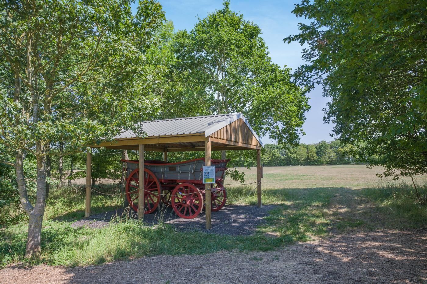 A covered wagon with red wheels parked under a wooden shelter in a grassy field.