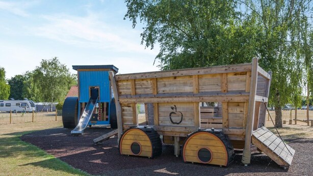 Un parque infantil con un módulo de juego de madera, que incluye un tobogán y una casita.
