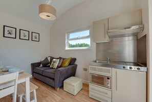 A compact kitchen with a built-in oven and microwave, a dining area with a table and chairs, a sofa with cushions, and a window with a view of greenery. - 1 Bed in Banham (oc-c1899) (Banham)