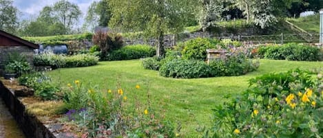 Un jardin où l’on trouve une grande variété de plantes et de fleurs, un mur de pierre et un bâtiment en arrière-plan.