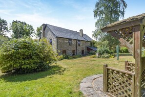 A stone building with a thatched roof, surrounded by a well-maintained lawn and a wooden fence. - 4 Bed in Newchurch (oc-bow333) (Newchurch)