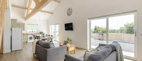 A spacious living area with a kitchen in the background, a large window, and a flat-screen TV mounted on the wall.