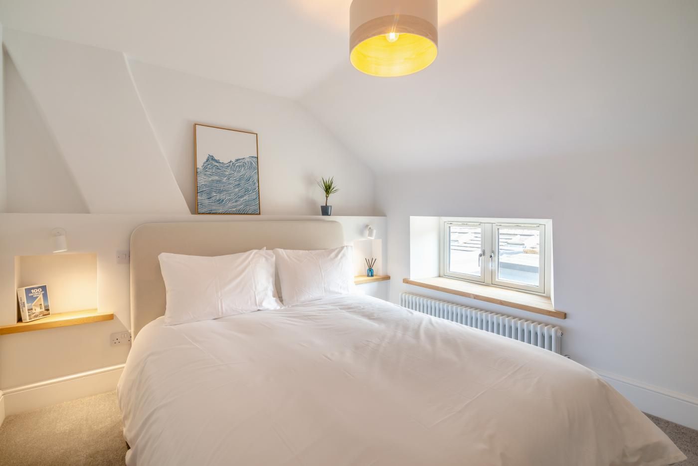 A bedroom with a bed, a bedside table, a window, a radiator, a wall-mounted light fixture, and a framed picture.