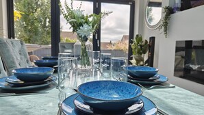 Una mesa de comedor con platos azules, vasos transparentes, un florero con follaje y vistas al exterior a través de amplias ventanas.