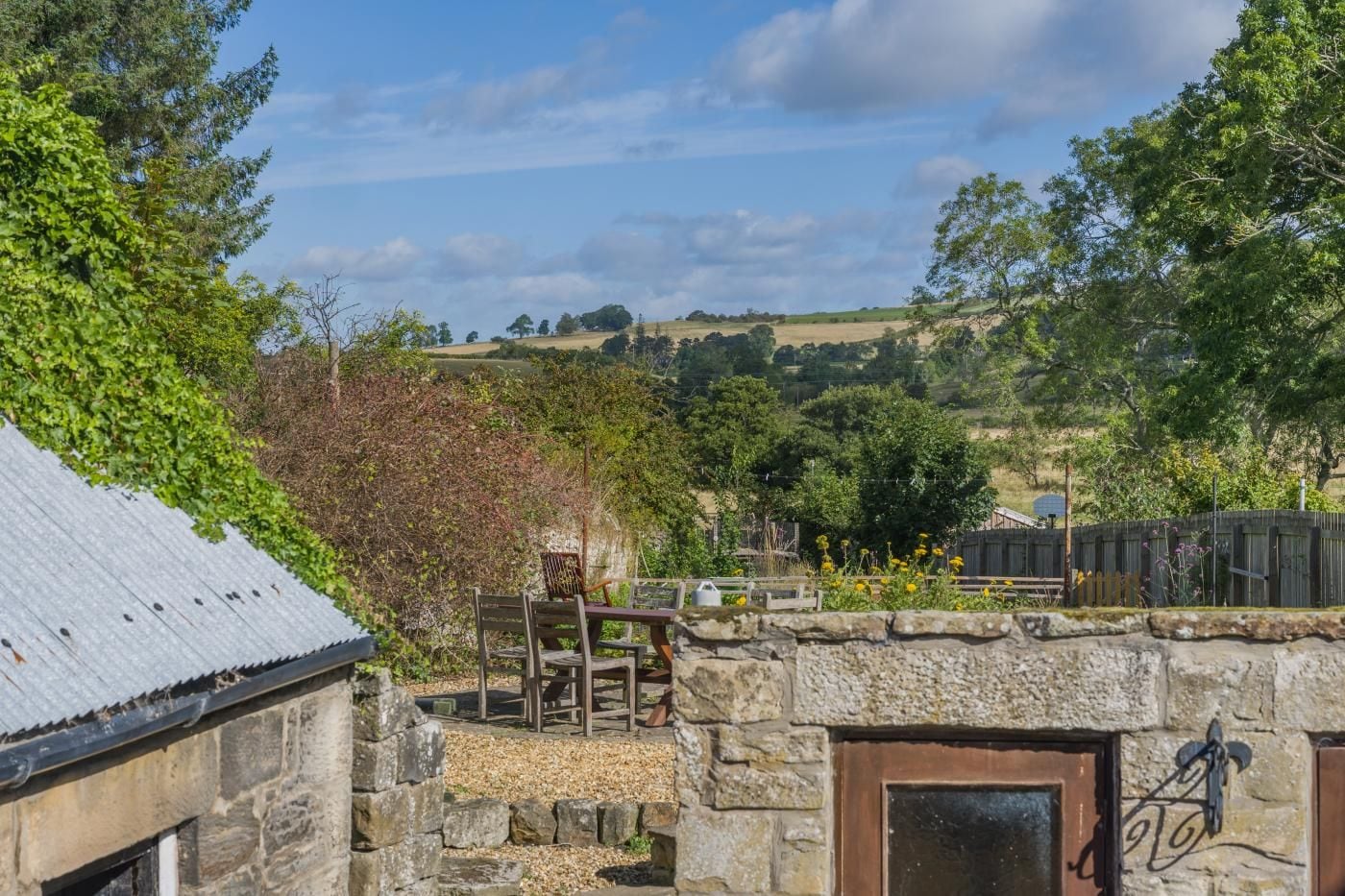 A stone building with a metal roof, a wooden fence, and a view of a hilly landscape.
