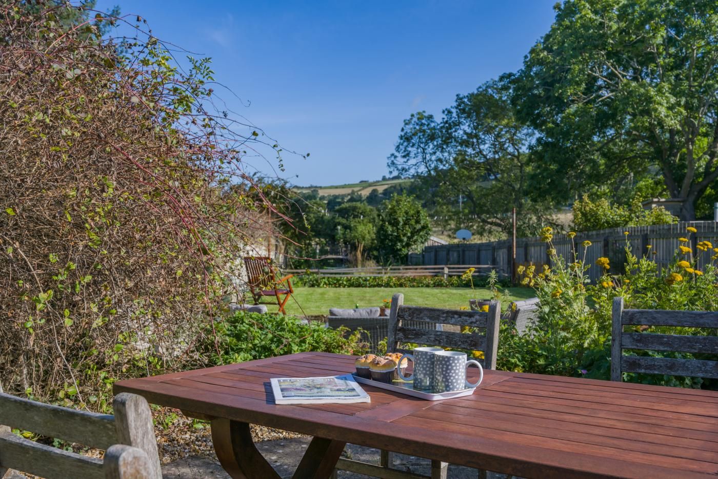 Ein Sitzbereich im Freien mit einem Holztisch und Stühlen, einer Bank und Blick auf einen Garten.