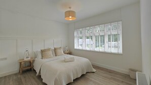 Une chambre à coucher avec un grand lit, une table de chevet, une commode, un luminaire suspendu et une fenêtre avec des stores.