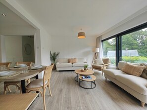Ein geräumiger Wohnbereich mit Esstisch, Sofa und Couchtisch, mit einem großen Fenster und Blick ins Grüne.