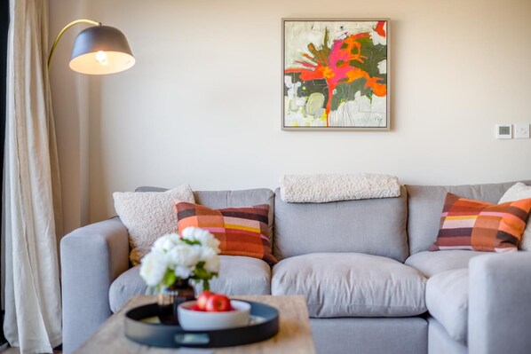 A modern living room with a grey sofa, colorful cushions, a white vase with flowers, and a painting on the wall.