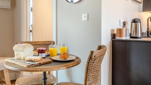 Une table ronde dressée pour le petit-déjeuner, avec du pain, des croissants et du jus, entourée de chaises en osier, et un comptoir de cuisine agrémenté d’une bouilloire et d’une machine à café.