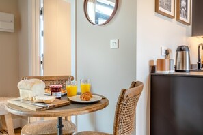 Ein Frühstückstisch mit Brot, Croissants und Saft, umgeben von Korbstühlen, mit einer Küchenzeile, die einen Wasserkocher und eine Kaffeemaschine enthält.