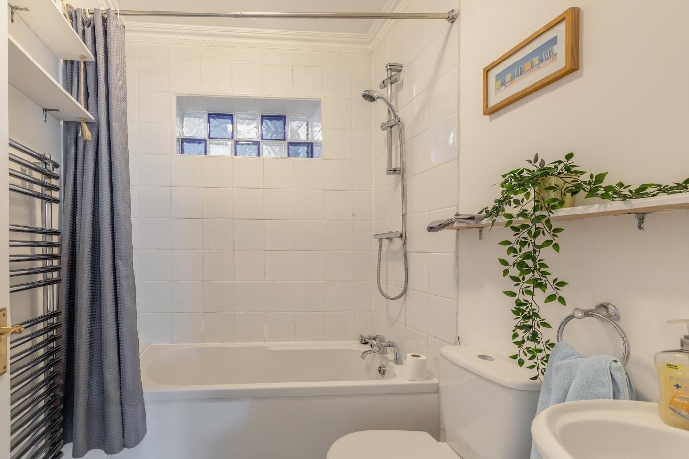 Casa de banho com banheira, chuveiro, sanita e lavatório. Tem uma janela com vidro azul e uma planta na parede.