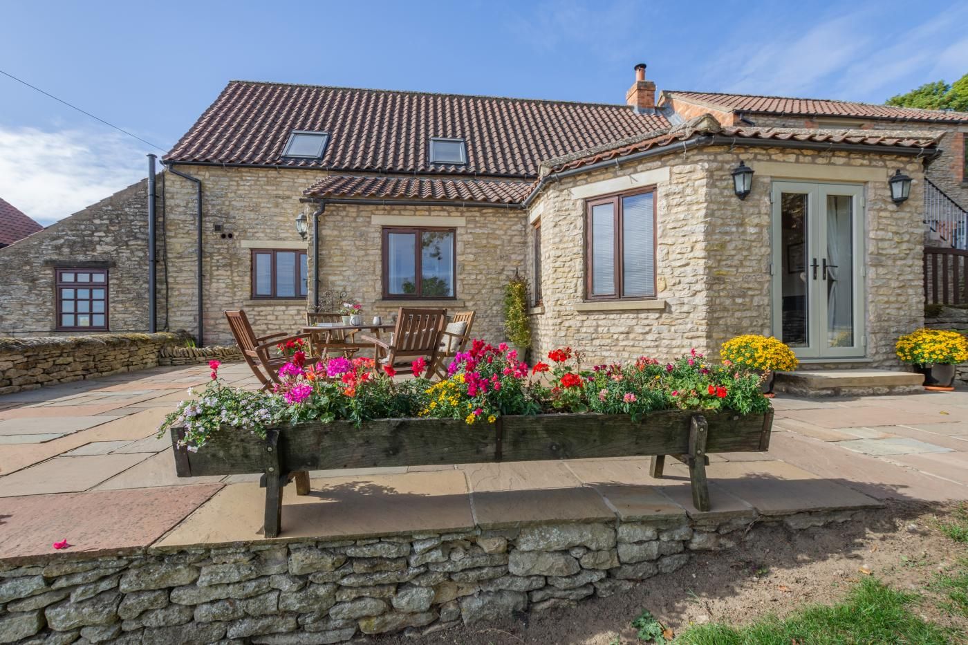 A stone house with a tiled roof, a patio with a wooden bench, and flower beds.