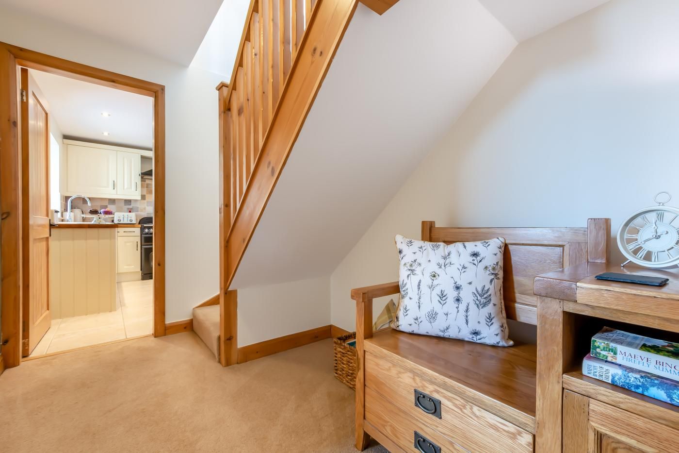 Een slaapkamer met een houten bed, een nachtkastje met boeken, een wandklok en uitzicht op een keuken en badkamer door een open deur.