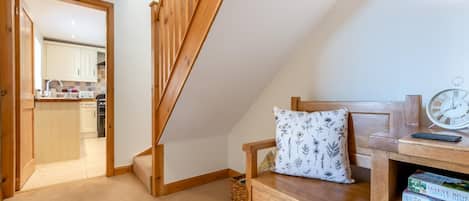 Un dormitorio con cama de madera, una mesita de noche con libros, un reloj de pared y vistas a una cocina y un baño a través de una puerta abierta.
