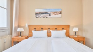A hotel room with a wooden headboard, two beds, two bedside tables with lamps, a window with curtains, and a beach scene painting on the wall. - Schone Ferienwohnung mit Wanne und Grossem Balkon in Sellin (Sellin)