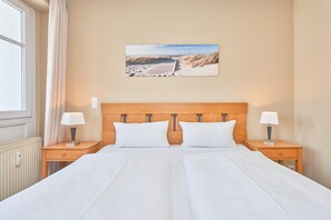 A hotel room with a wooden headboard, two beds, two bedside tables with lamps, a window with curtains, and a beach scene painting on the wall. - Schone Ferienwohnung mit Wanne und Grossem Balkon in Sellin (Sellin)