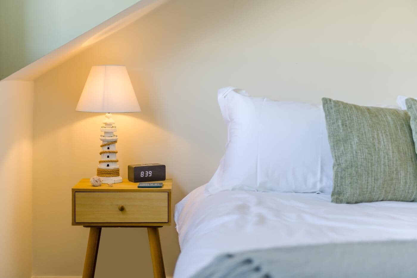 A neatly made bed with white bedding and a green throw pillow, a bedside lamp, and a digital clock on a wooden bedside table.