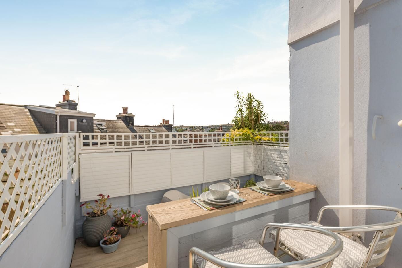 A balcony with a wooden table set for two, surrounded by potted plants and a white fence.