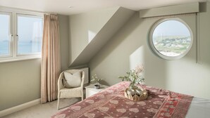 Una camera da letto con un letto, una sedia, un tavolino, un vaso con fiori, una finestra con tende e vista sul mare.
