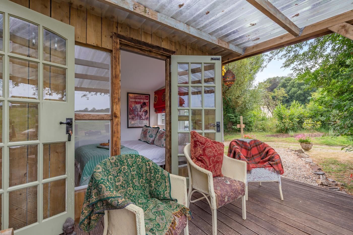 An open wooden room with a bed, two chairs, and a view of a garden.