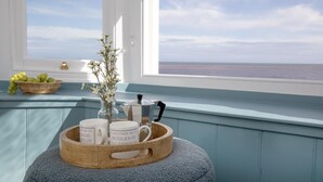 A view from a window with a wooden tray holding mugs and a kettle, a vase with flowers, and a basket of grapes on a blue cushioned bench. - 3 Bed in Budleigh Salterton (oc-a44975) (Budleigh Salterton)