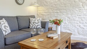Uma sala de estar com um sofá cinzento, uma mesa de centro de madeira com taças de vinho e um vaso de flores, um candeeiro e um relógio na parede.