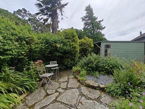 A stone patio with a table and chairs, surrounded by lush greenery and a small shed. - 2 Bed in Kingston (oc-s43307) (Kingston)