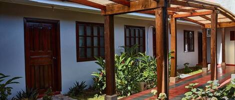 A covered outdoor area with wooden beams, a stone pathway, and a building with a wooden door and windows.