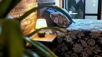 A room with a brick wall, a bed with patterned bedding, a bedside table with a lamp, and a view through a glass door.
