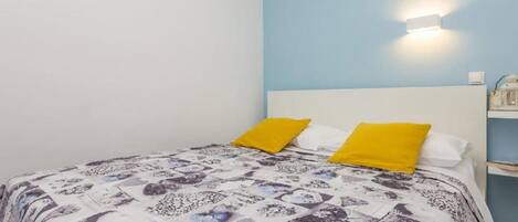 A bedroom with a bed, two pillows, a wall-mounted light, and a nightstand with a lamp.