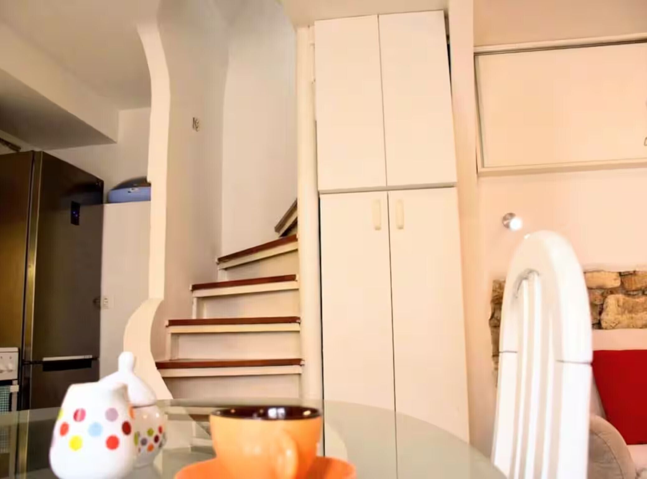 Una cocina con un armario blanco, una escalera de madera y una mesa de vidrio con tetera y taza.