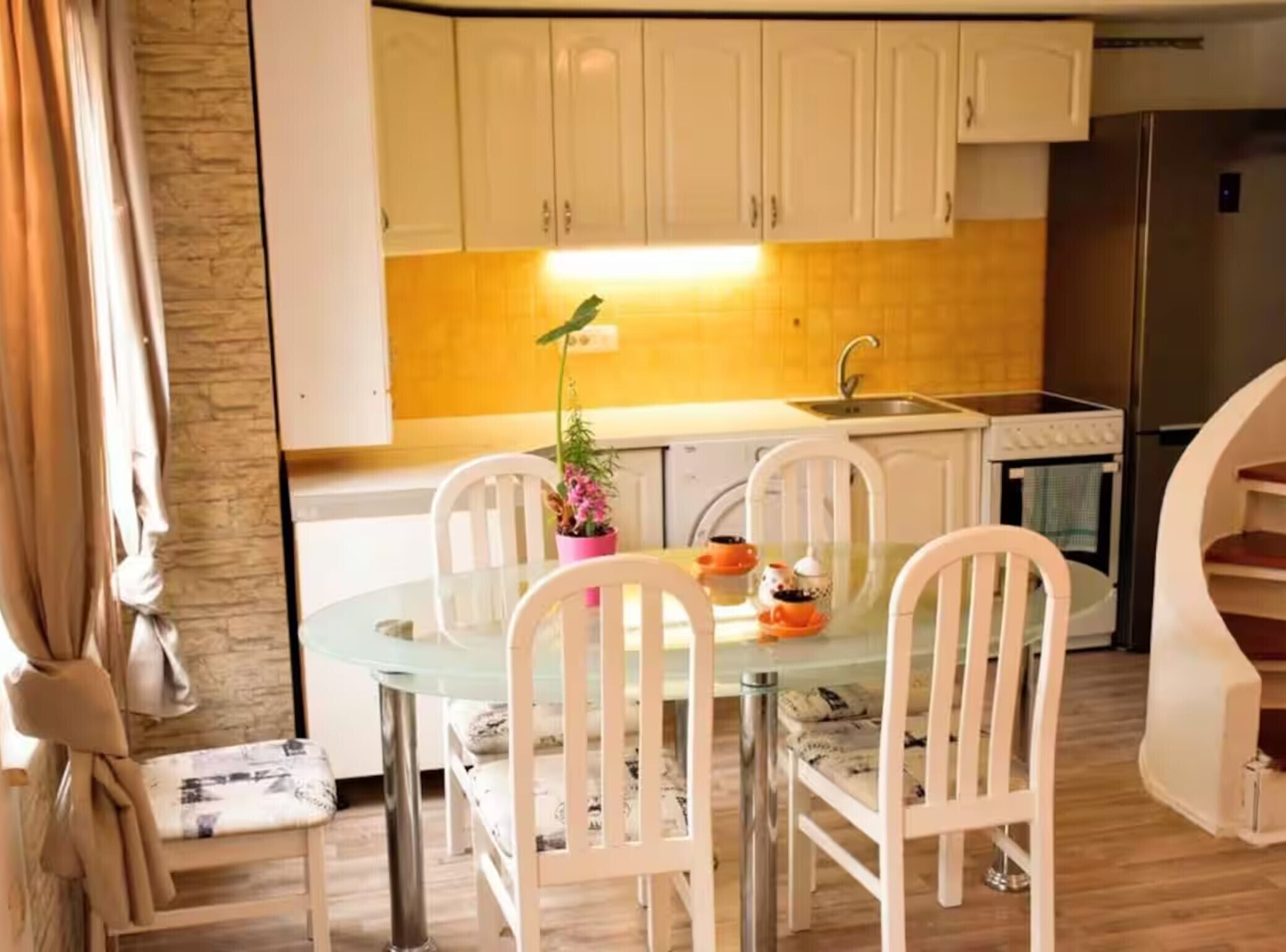 Une cuisine compacte avec des meubles blancs, un réfrigérateur en acier inoxydable, une table à manger en verre avec des chaises blanches et une plante en pot.