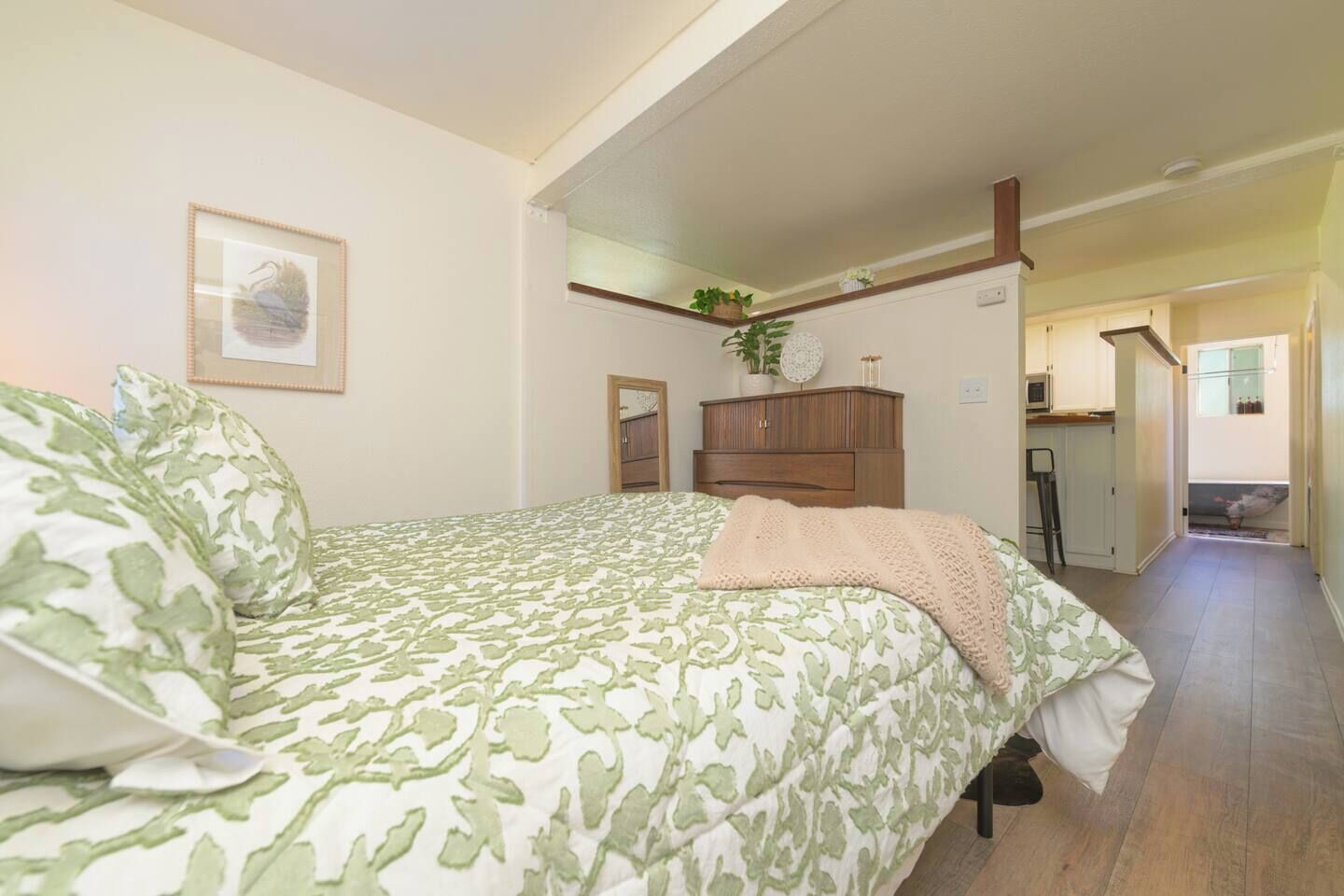 Un dormitorio con cama, cómoda, espejo y vista a la cocina.