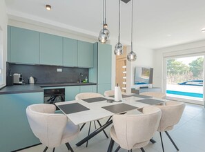 A modern kitchen with teal cabinets, a dining area with a table and chairs, and a large window with a view of a boat. - Three Bedroom Villa with Outdoor Swimming Pool (Vodice)