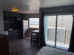 A compact room with a kitchenette, a bed, a small table, and a view of the sea through a large window. - View 2 Queen South -PetOK-Studio-Balcony-Tradition (Myrtle Beach)