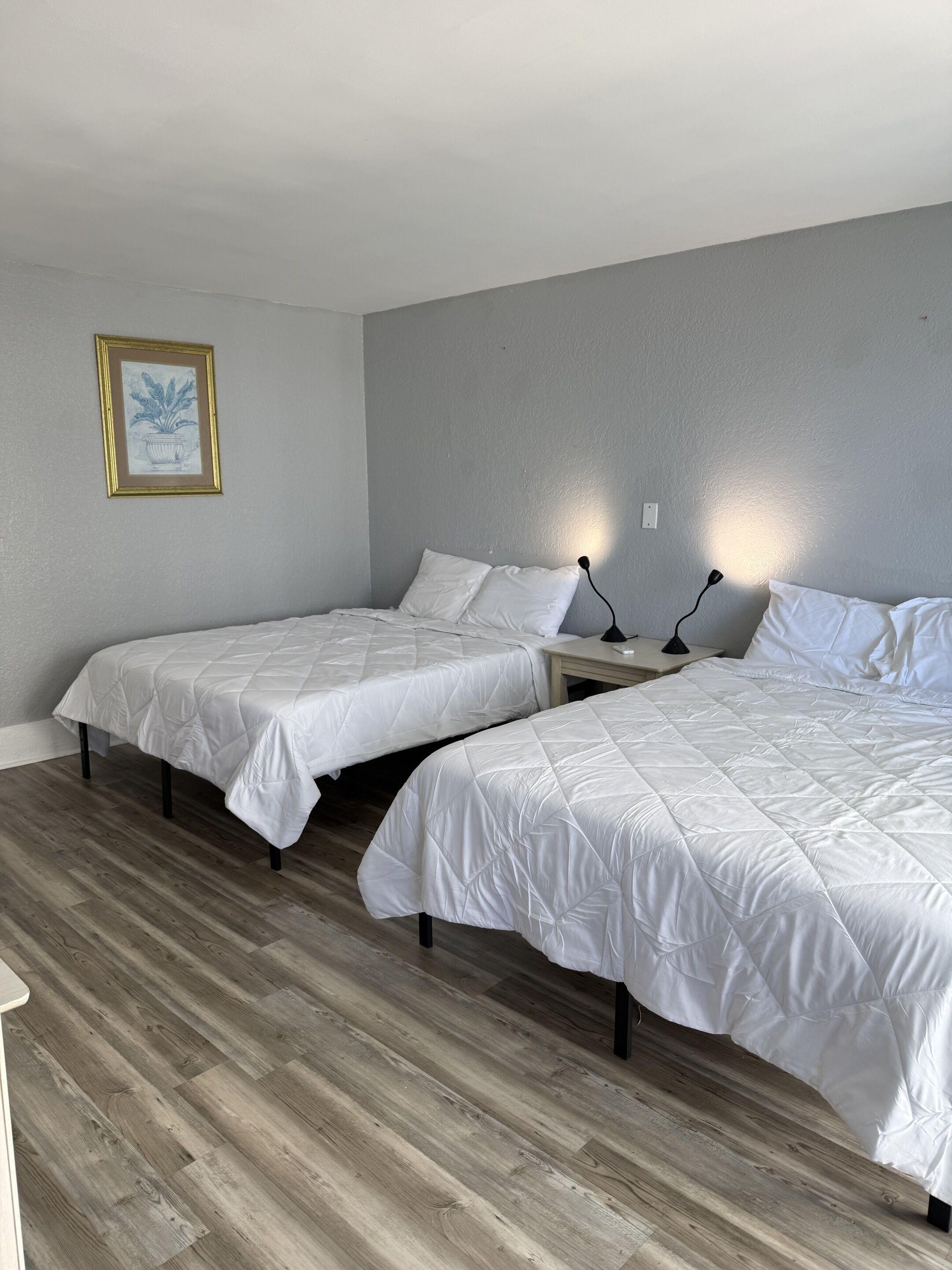 A room with two beds, a bedside table with lamps, a framed picture on the wall, and wooden flooring.