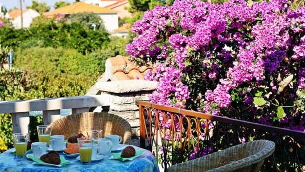 Ein Balkon mit Korbmöbeln, einem Tisch, der mit Frühstück zubereitet ist, und einem großen Busch mit violetten Blüten.