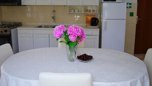 Et køkken med hvide skabe, en mikrobølgeovn og en vask. Der er et bord med hvid dug, en vase med lyserøde blomster og en skål med kirsebær.