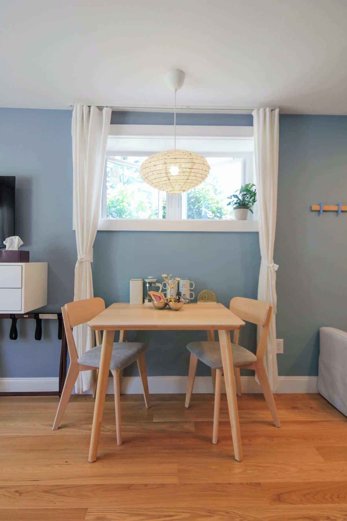Un comedor con una mesa y sillas, una lámpara colgante, una ventana con cortinas y una planta en el alféizar.