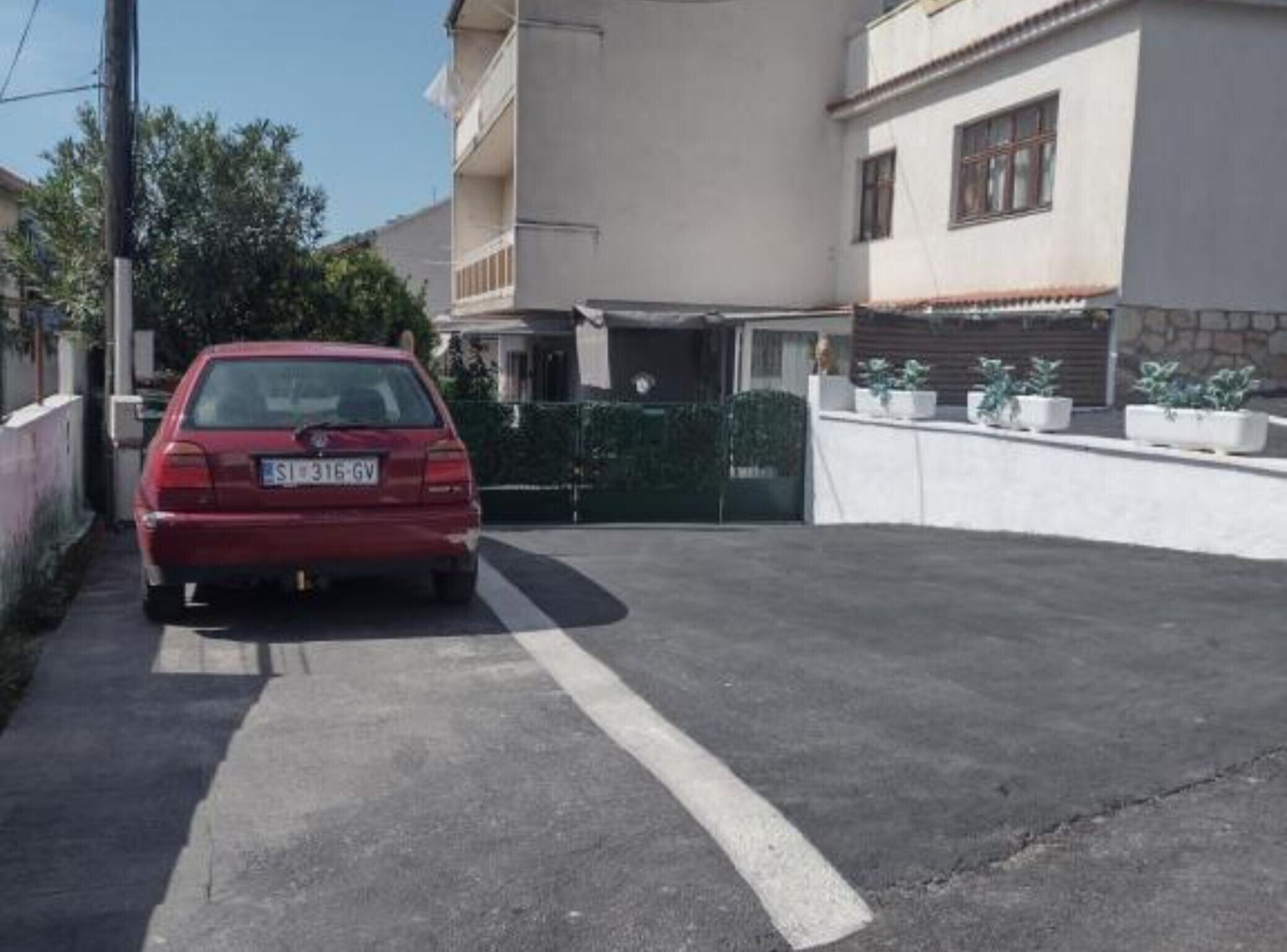 Une voiture rouge est garée dans une allée, délimitée par une ligne blanche, avec une entrée fermée par un portail et un bâtiment doté d’une façade en pierre et de fenêtres.