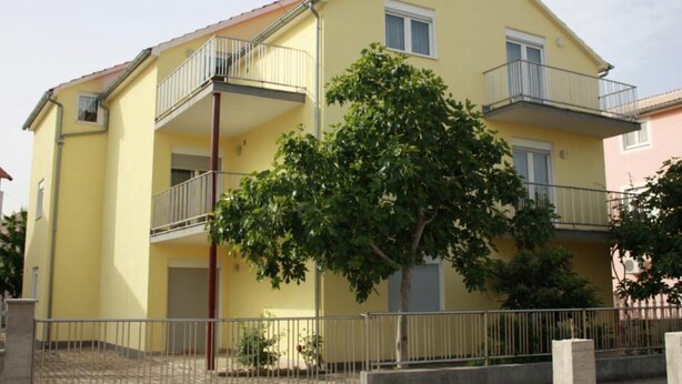Ein gelbes, zweistöckiges Gebäude mit Balkonen, einem Baum davor und einem Metallzaun.