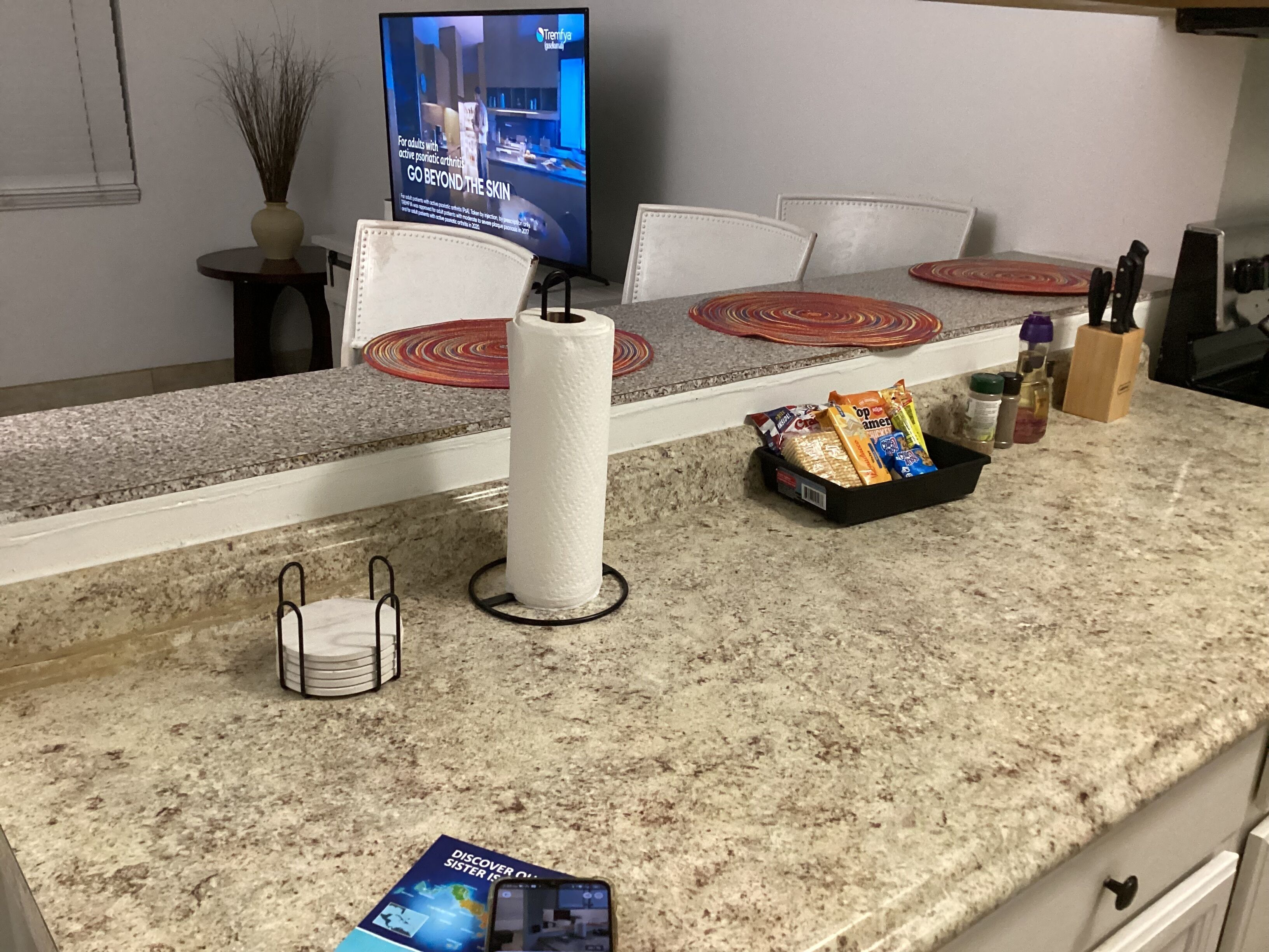 A kitchen counter with a television, a paper towel holder, a bread basket, and a magazine.