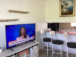 Un televisor mostrando un noticiero, una zona de comedor con sillas, un área de cocina y un cuadro en la pared.