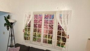 Una habitación con ventana que ofrece vistas a un jardín floral, un sofá oscuro con cojines y una lámpara de pie.
