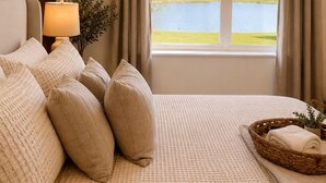 A bedroom with a large window, a bed with multiple pillows, a bedside lamp, and a wicker tray with a towel.