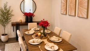 A dining area with a wooden table, white chairs, a vase with red flowers, and three wall-mounted abstract paintings.