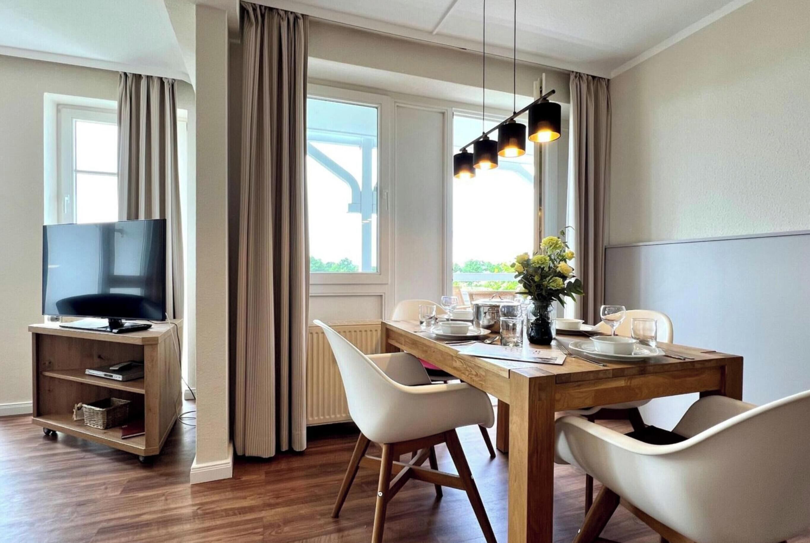 A dining area with a wooden table, white chairs, a TV on a stand, and a window with curtains.