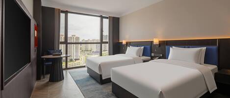 A hotel room with two beds, a desk, and a large window offering a city view.